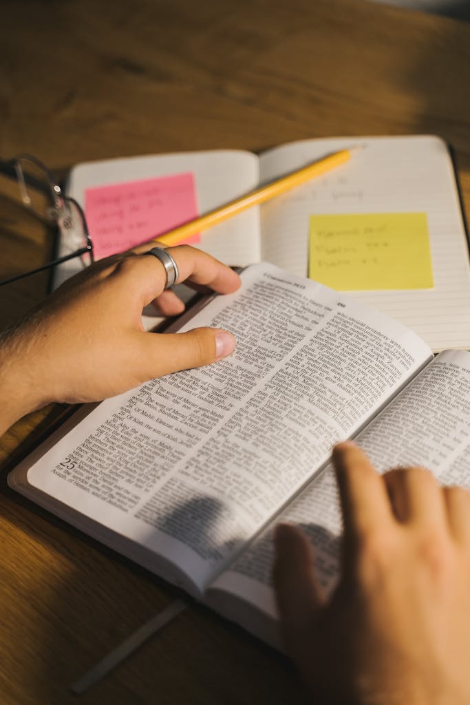 Close-up of hands holding and reading a Bible with study notes and a pencil on a desk.