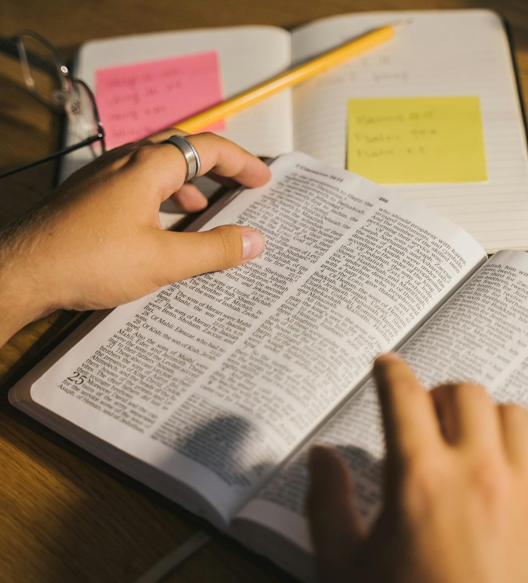 Close-up of hands holding and reading a Bible with study notes and a pencil on a desk.