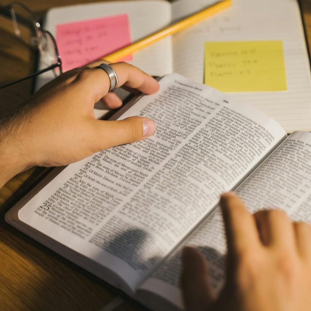 Close-up of hands holding and reading a Bible with study notes and a pencil on a desk.
